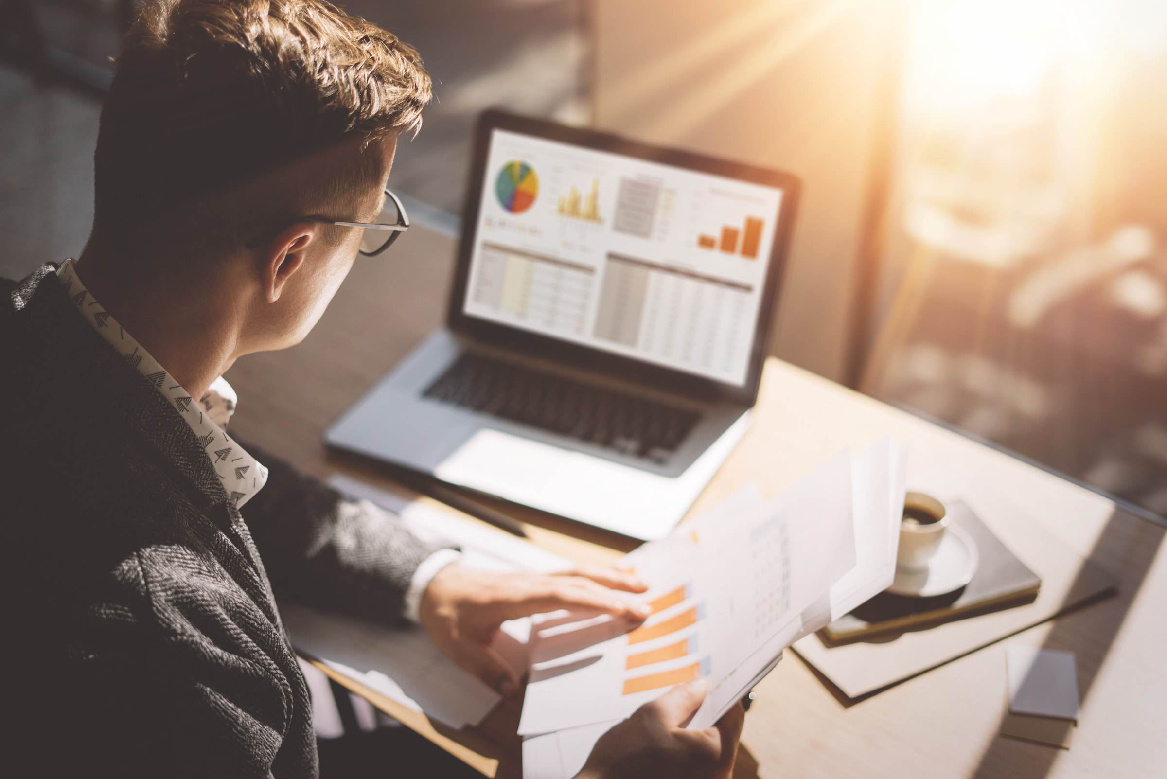 man looking at financial graphs papers spreadsheets on laptop in amber light